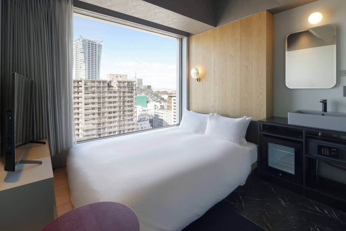 A hotel room with a large bed, a wooden wall, a modern bathroom, and a view of the cityscape through the window.