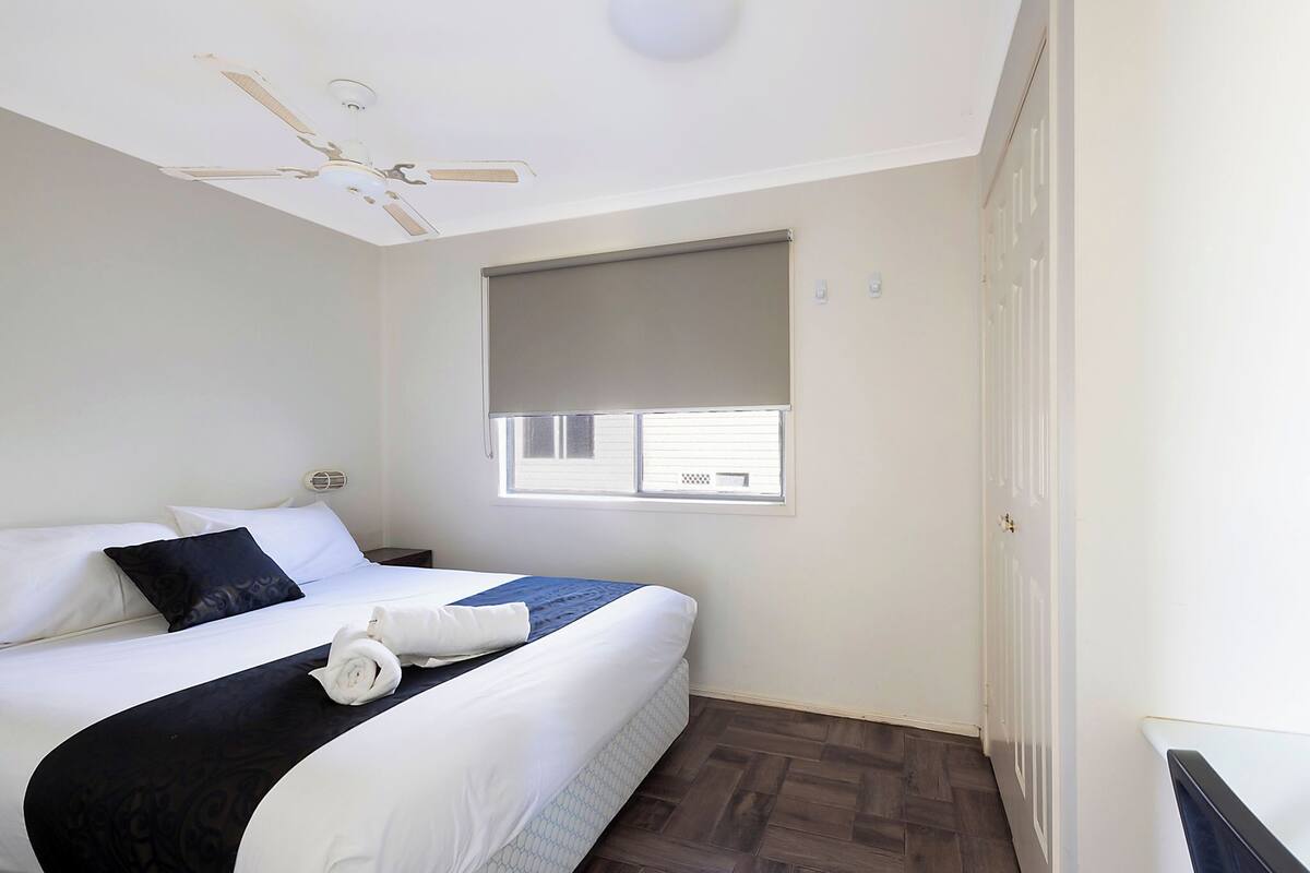 A hotel room with a bed, a ceiling fan, a window with blinds, and a door.