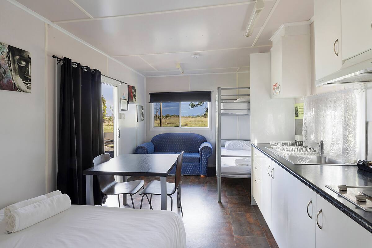 A compact room with a bed, a table, a chair, a sofa, a window with a view, and a kitchenette.