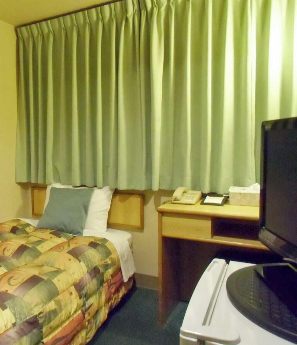 Habitación de hotel con cama, escritorio con teléfono y papeles, televisor y un ventanal con cortinas.