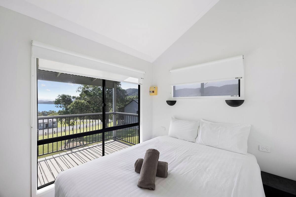 A bedroom with a bed, white bedding, a balcony with a view of the outdoors, and a wall-mounted TV.