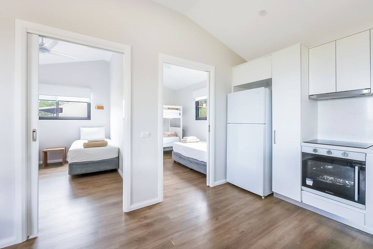 A modern kitchen with white cabinets and a built-in oven. There are two bedrooms visible, one with a bed and another with a bunk bed.