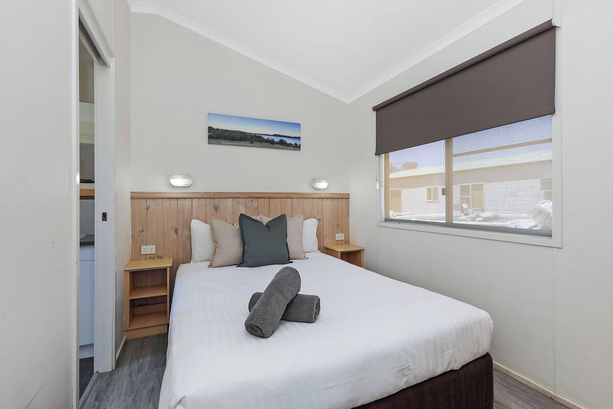 A neatly made bed with a white duvet, grey towels, and a wooden headboard. There is a window with a brown blind, a wall-mounted picture, and a small bedside table with a lamp.