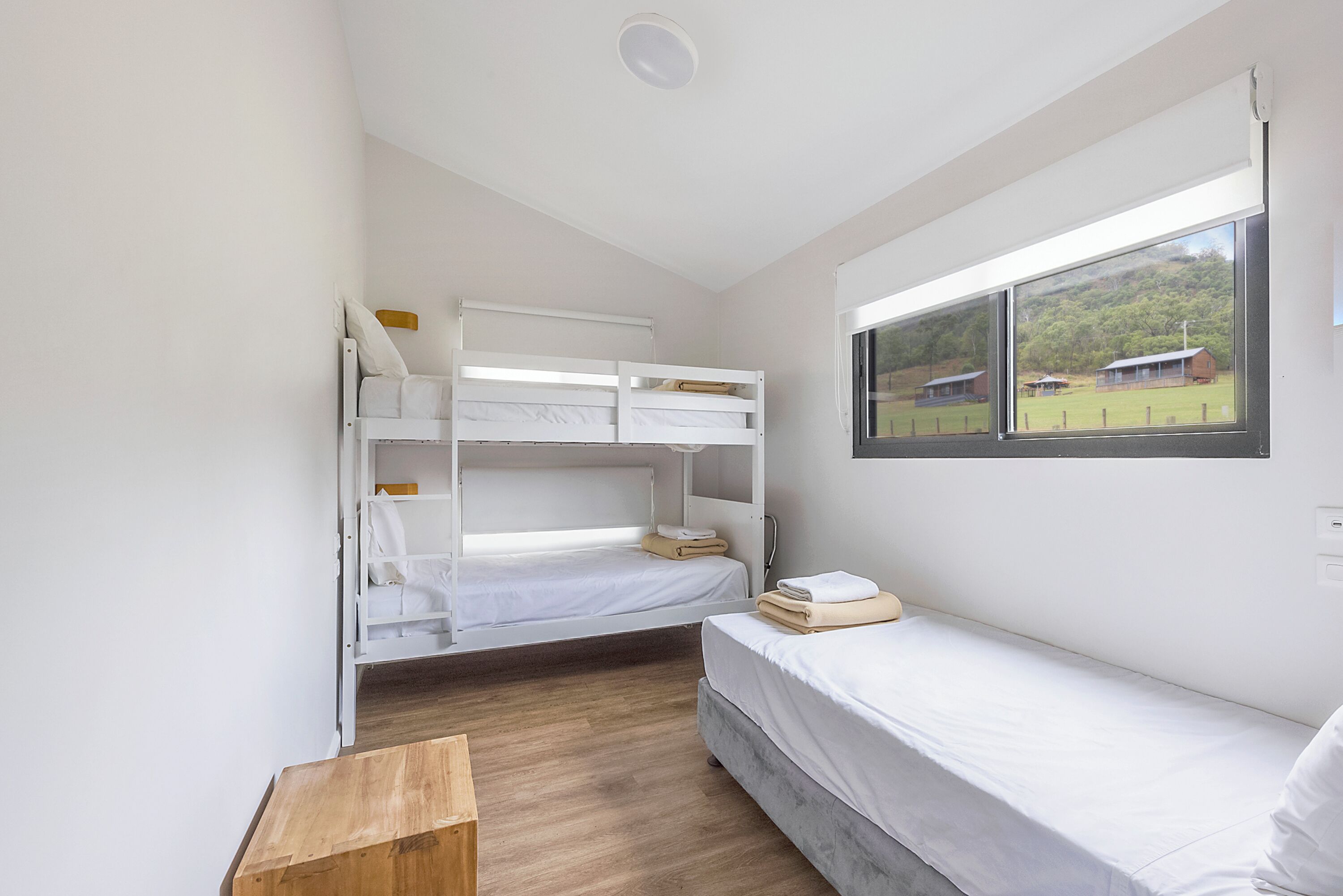 A room with bunk beds, a single bed, a wooden table, and a window with a view of a house and trees.