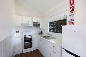 A modern kitchen with white cabinets, a stainless steel microwave, a stove, and a sink. There are fire safety signs on the wall. - Reflections Lake Glenbawn - Holiday Park (Glenbawn)
