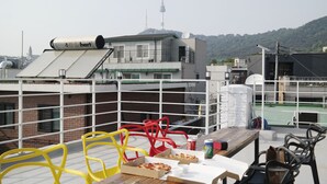 Eine Dachterrasse mit Tisch, Stühlen, Pizzakartons und einer Limodose. Im Hintergrund ist ein großes Gebäude mit einem Turm zu sehen.
