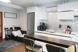 A compact kitchen with white cabinets, a stainless steel refrigerator, a microwave, and a coffee maker. There is a small dining area with a wooden table and two chairs.