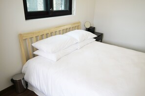 A neatly made bed with white linens, a wooden headboard, a small bedside table with a mirror, and a trash bin.