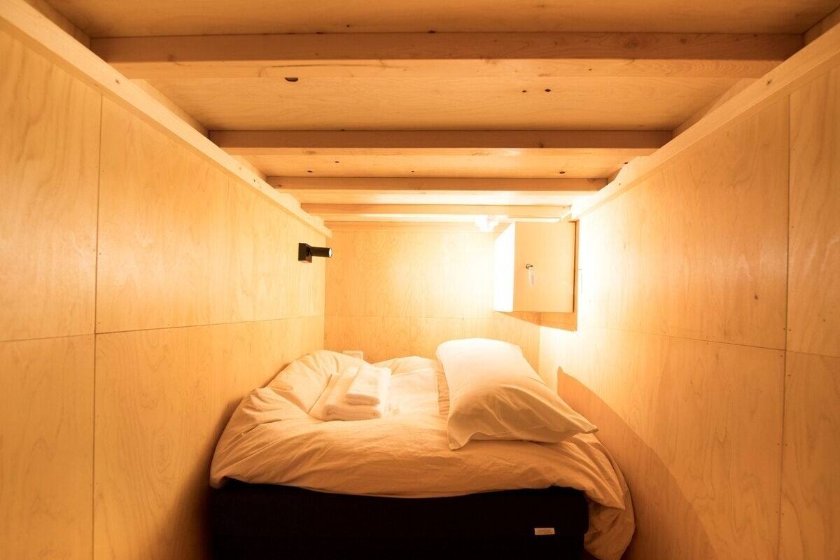 A small, enclosed wooden room with a bed, a wall-mounted light, and a small cabinet.
