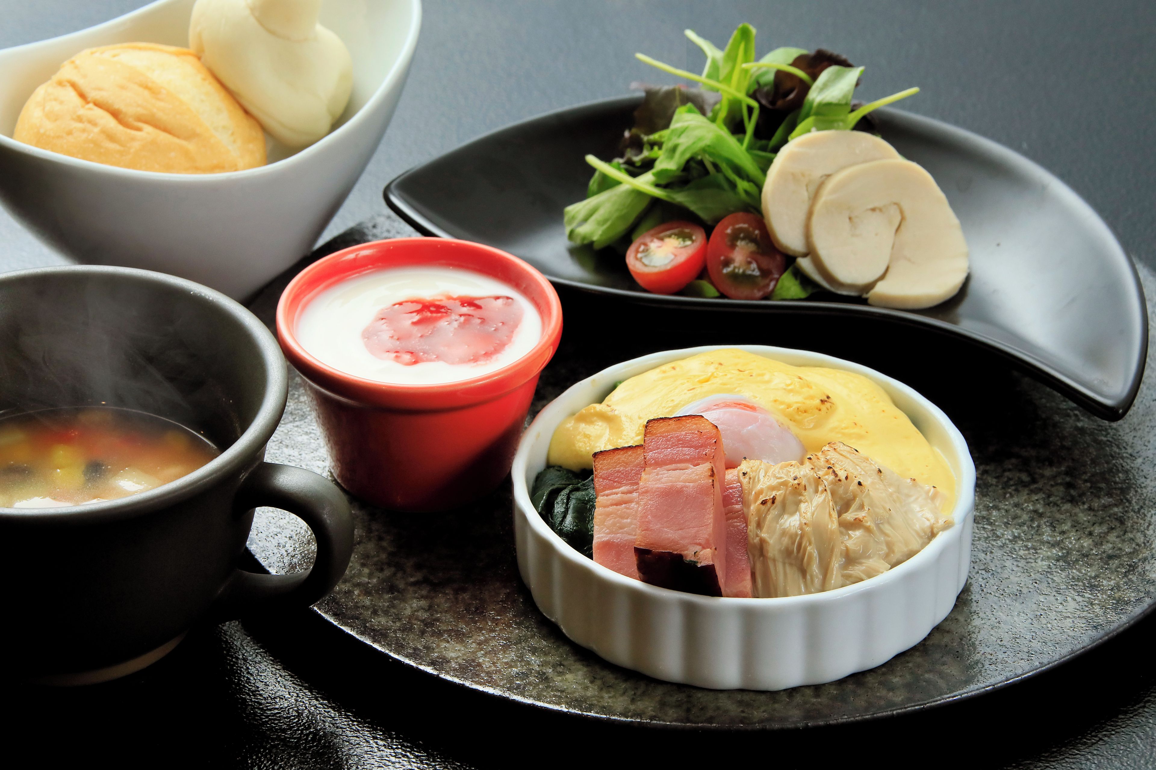 A meal with soup, a red bowl with a white substance, a plate with greens and sliced items, and a bowl with a yellow substance and meat.