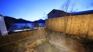 Un bain à remous en pierre, doté d’une terrasse en bois et entouré d’une clôture en bois, offrant une vue sur un lac et des montagnes.