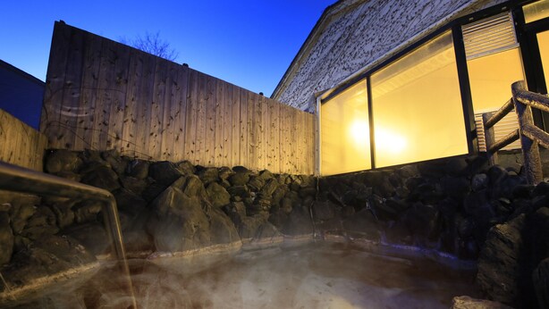 A hot spring area with a wooden fence, stone wall, and a wooden staircase leading to a building with large windows. - YUTORELO NIKKO - Hostel (Nikko)