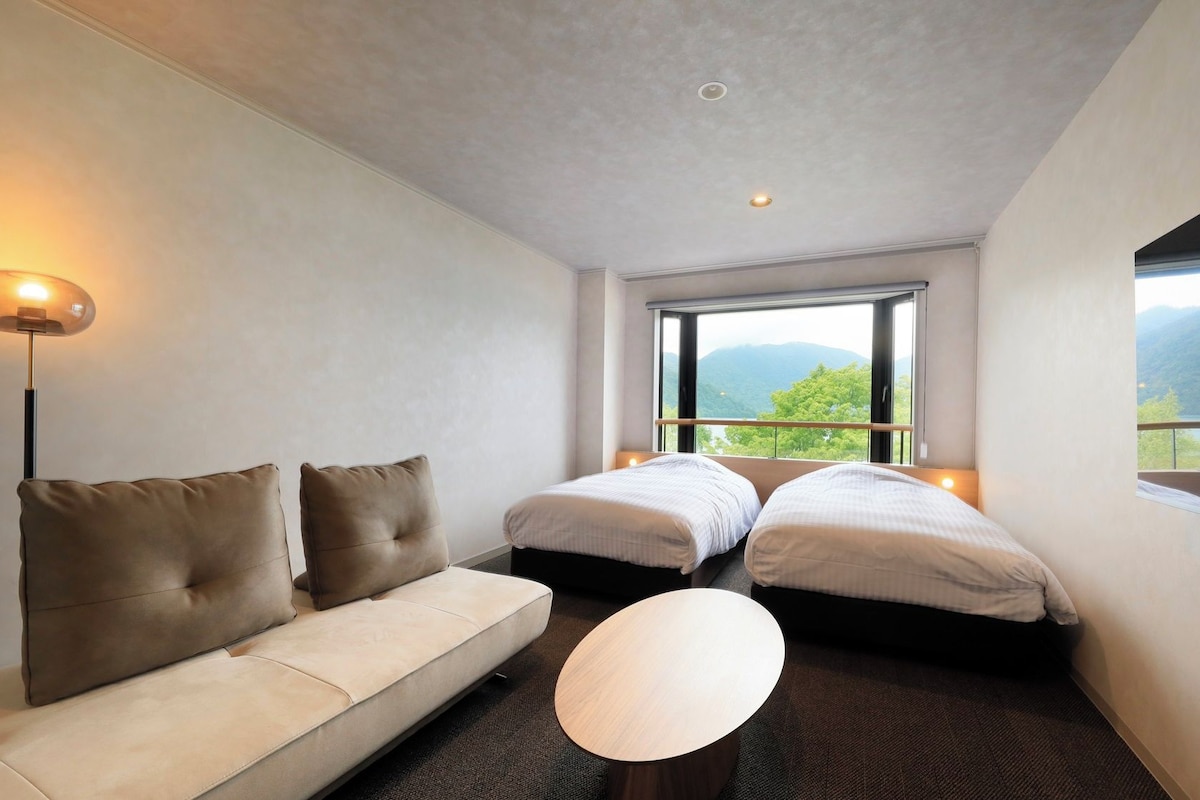 A hotel room with a sofa, two beds, a round table, and a large window with a mountain view.