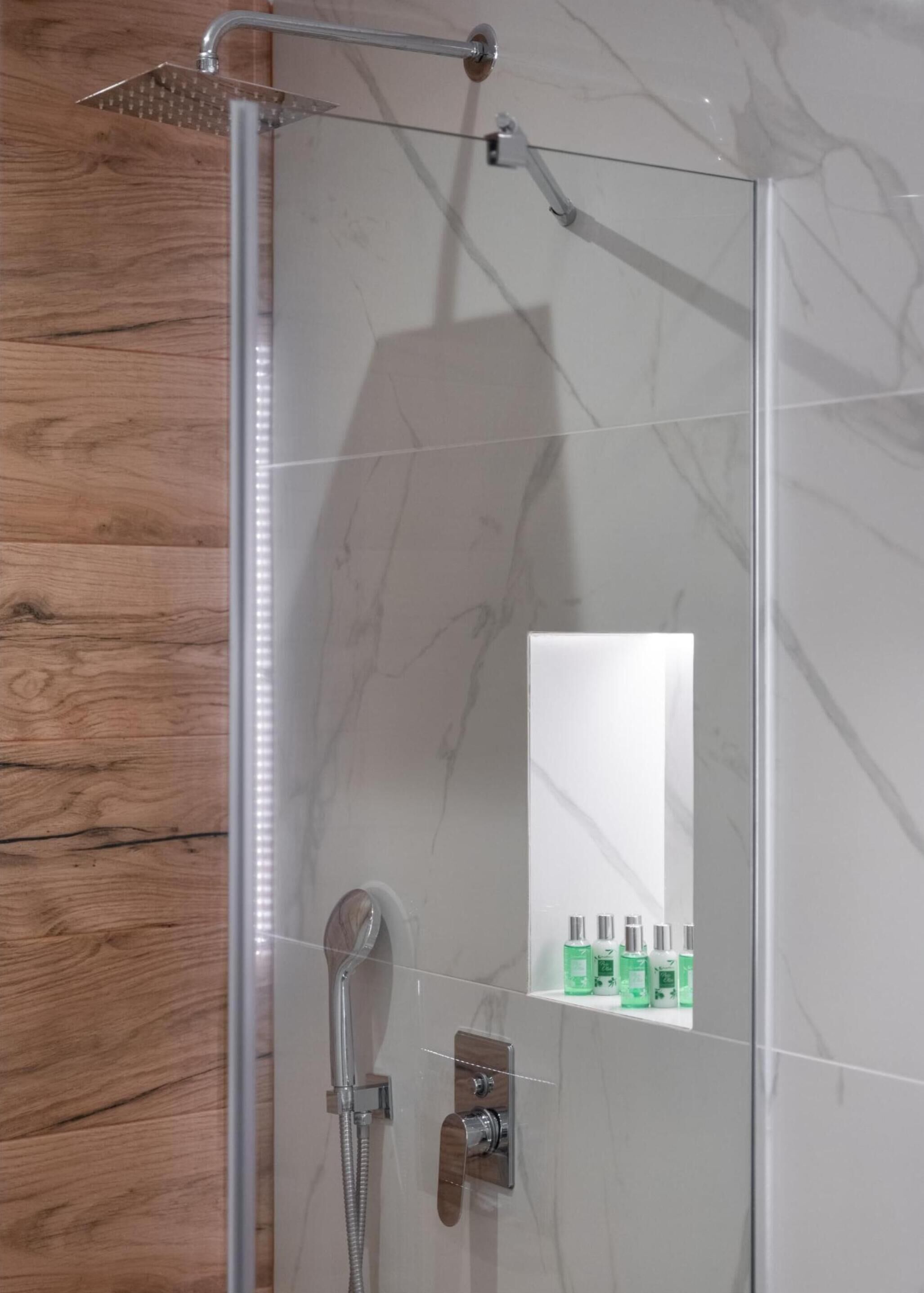 A modern shower with a handheld showerhead, a handheld shower attachment, and a wall-mounted control. There is a mirror above the shower, and a shelf with toiletries.