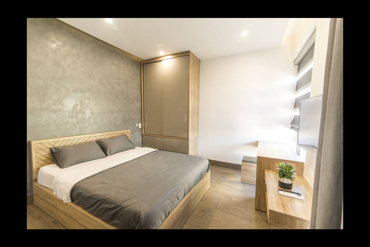 Un dormitorio moderno con una cama de madera, un colchón gris, cabecera gris, un armario, una mesita de noche con una planta y un televisor en la pared.