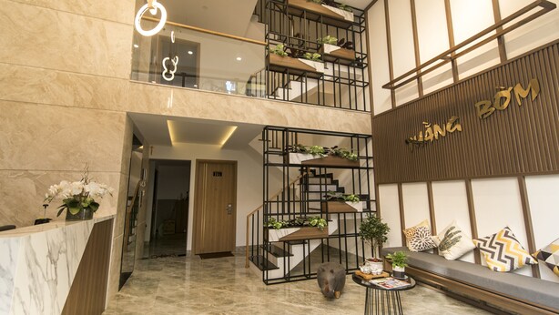 Un moderno vestíbulo de hotel con un mostrador de recepción de mármol, una escalera con plantas y una zona de estar con cojines.