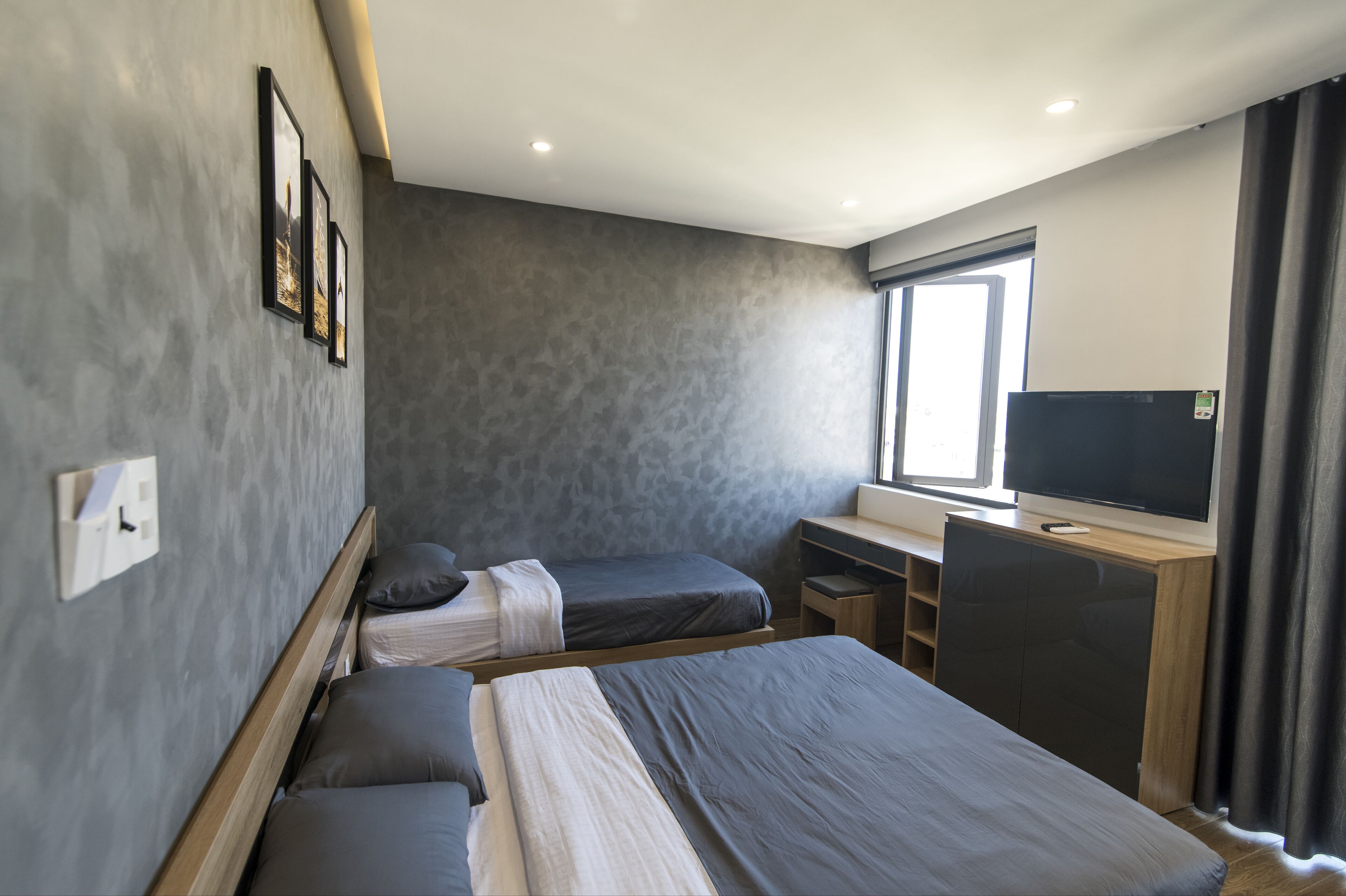 A modern hotel room with a large bed, two smaller beds, a desk with a TV, and a window with curtains.