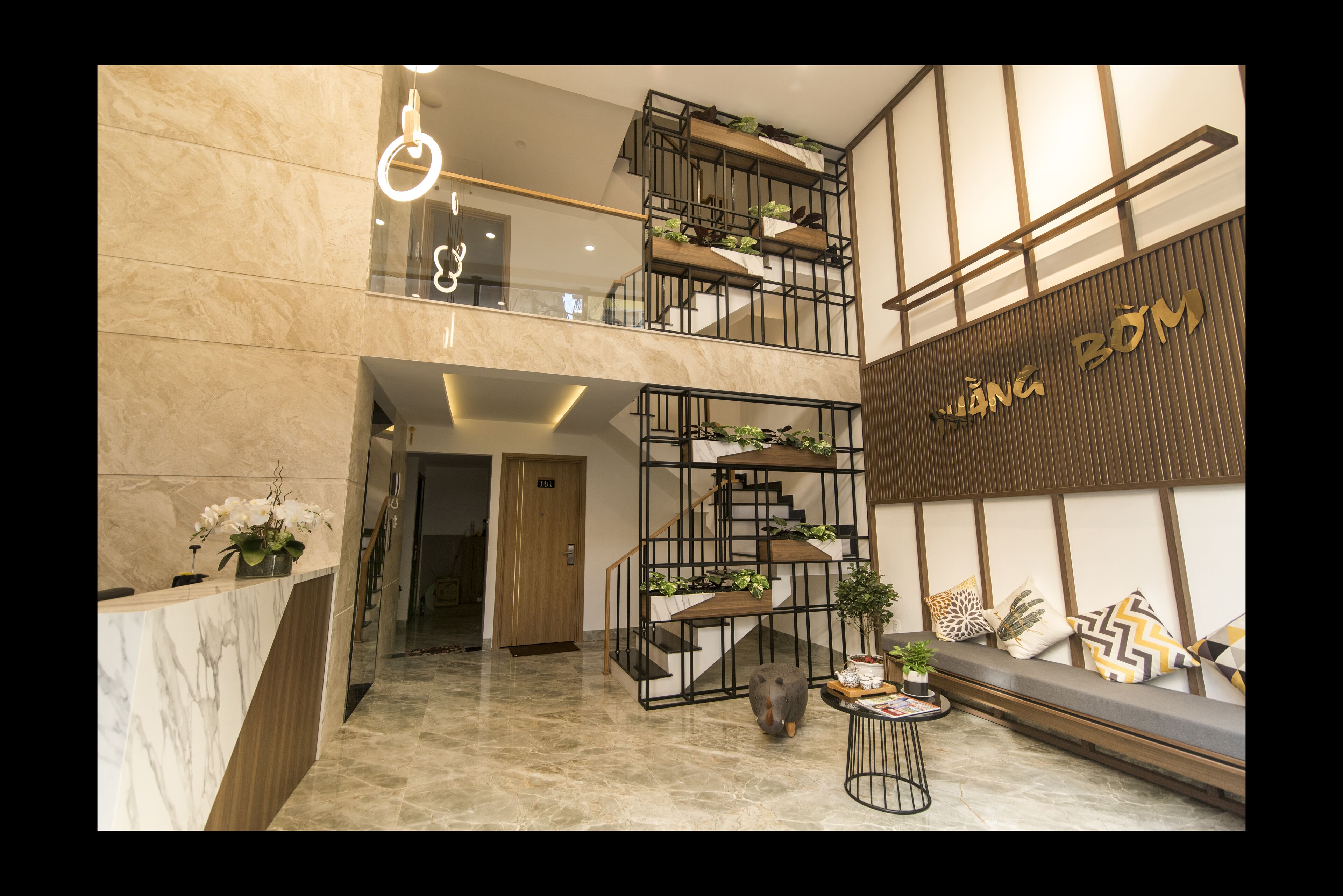A modern hotel lobby with a marble reception desk, a staircase with plants, and a seating area with decorative pillows.