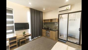 A modern hotel room with a bed, a desk with a TV, a kitchenette, and a sliding glass door to the bathroom.