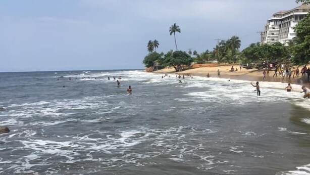 A beach with waves, palm trees, and a building in the background. - Hotel Samaki Plage (Kribi)