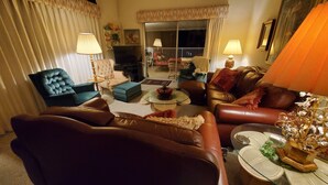 Una sala de estar con sofás de cuero, una mesa de centro de vidrio y lámparas. Hay un televisor y una ventana con cortinas.