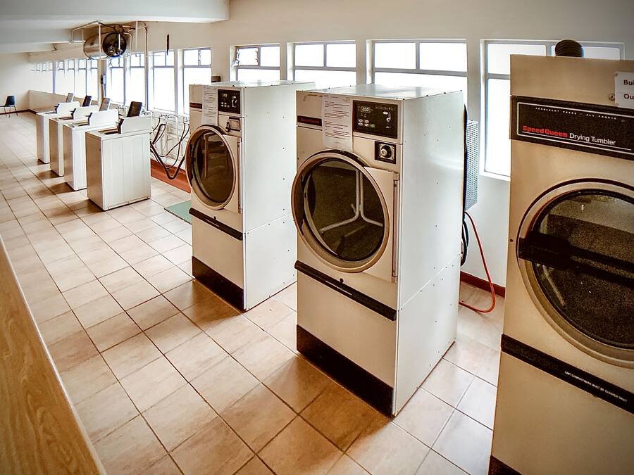 A row of front-loading washing machines in a well-lit laundry room with large windows.