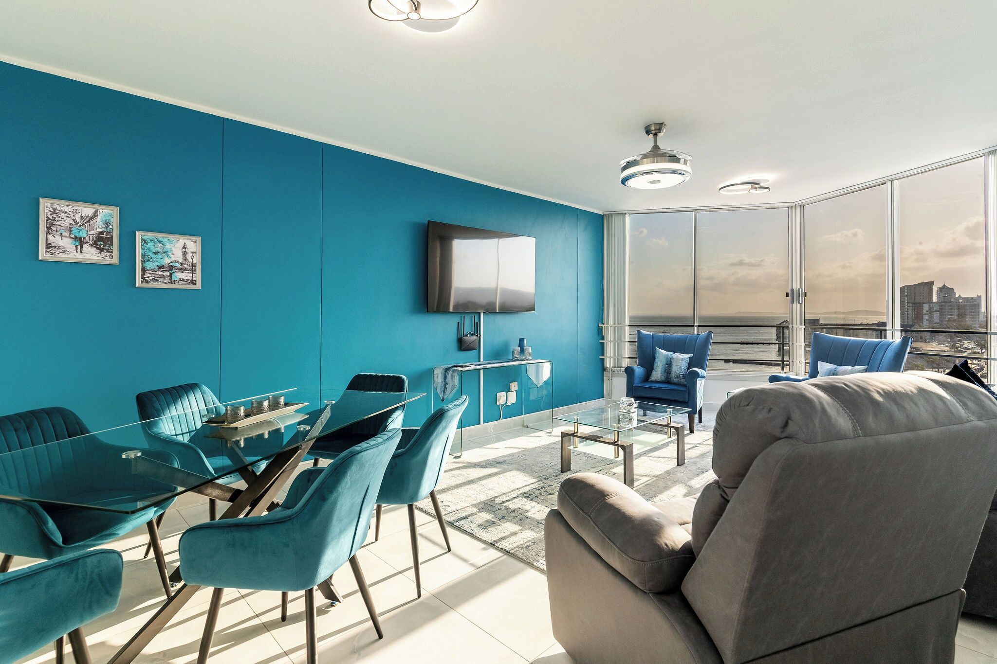 Un salon moderne avec un mur bleu, une table à manger en verre, des chaises bleues, un canapé gris et un téléviseur fixé au mur.
