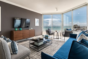 A modern living room with a large TV, a sofa, and a coffee table. There is a view of the ocean through large windows.
