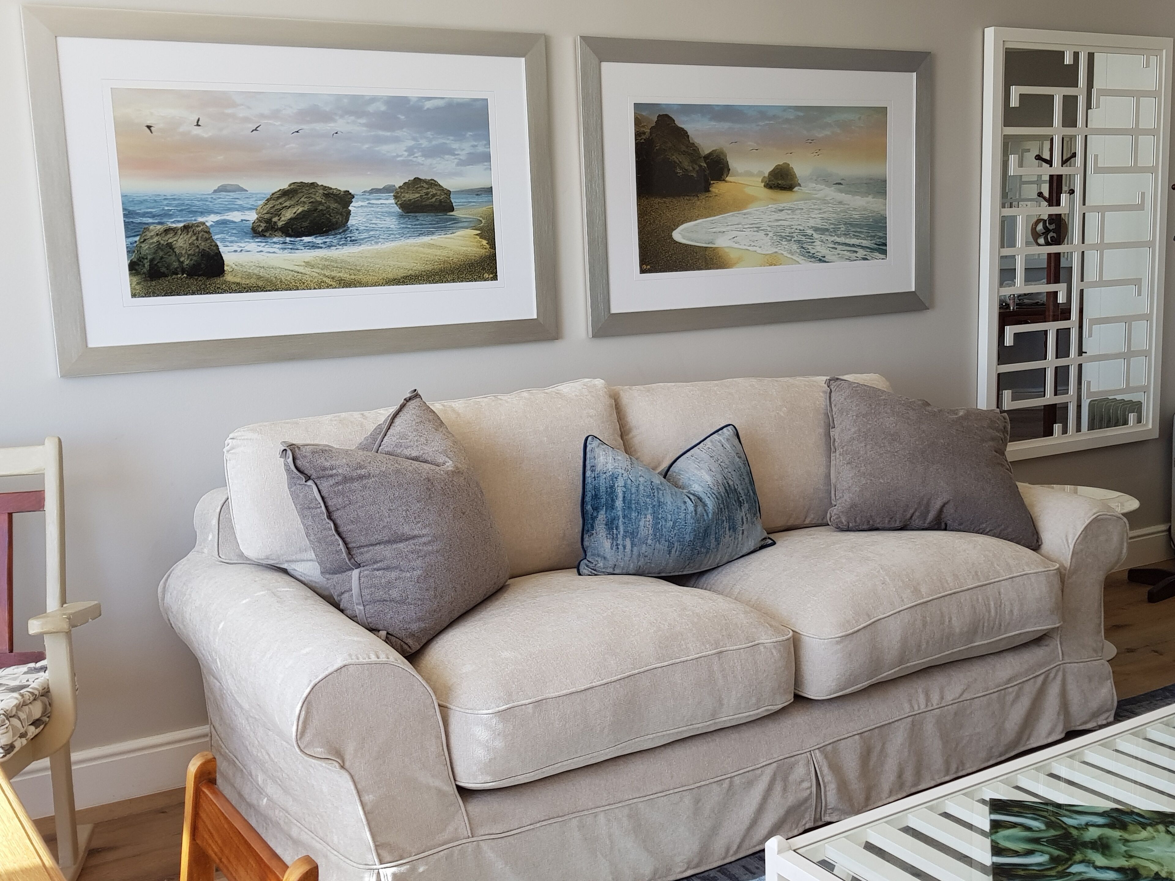 Een beige bank met drie kussens, twee ingelijste strandlandschappen aan de muur en een witte kast met een geometrisch ontwerp.