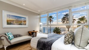 Ein Schlafzimmer mit großem Fenster mit Blick auf einen Strand, einem Bett mit vielen Kissen, einem Sofa mit Sitzkissen und einem gerahmten Bild an der Wand.