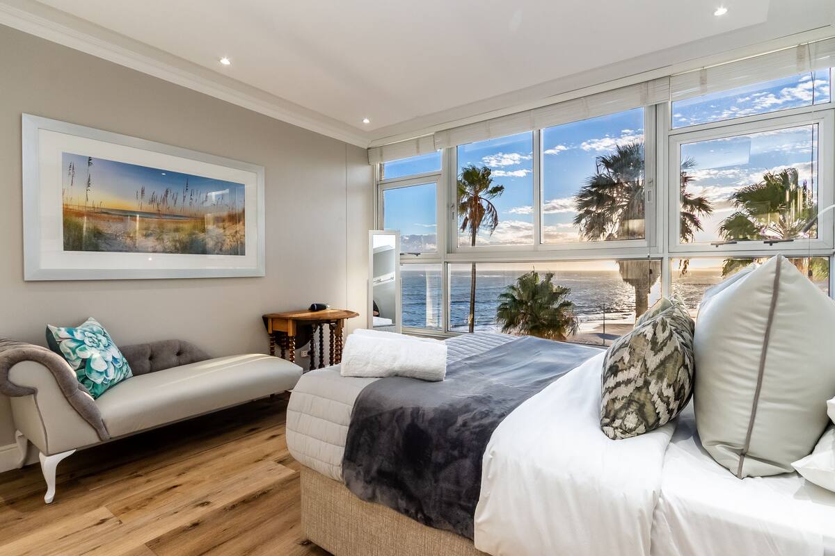 A bedroom with a large window overlooking a beach, a bed with multiple pillows, a sofa with a cushion, and a framed picture on the wall.