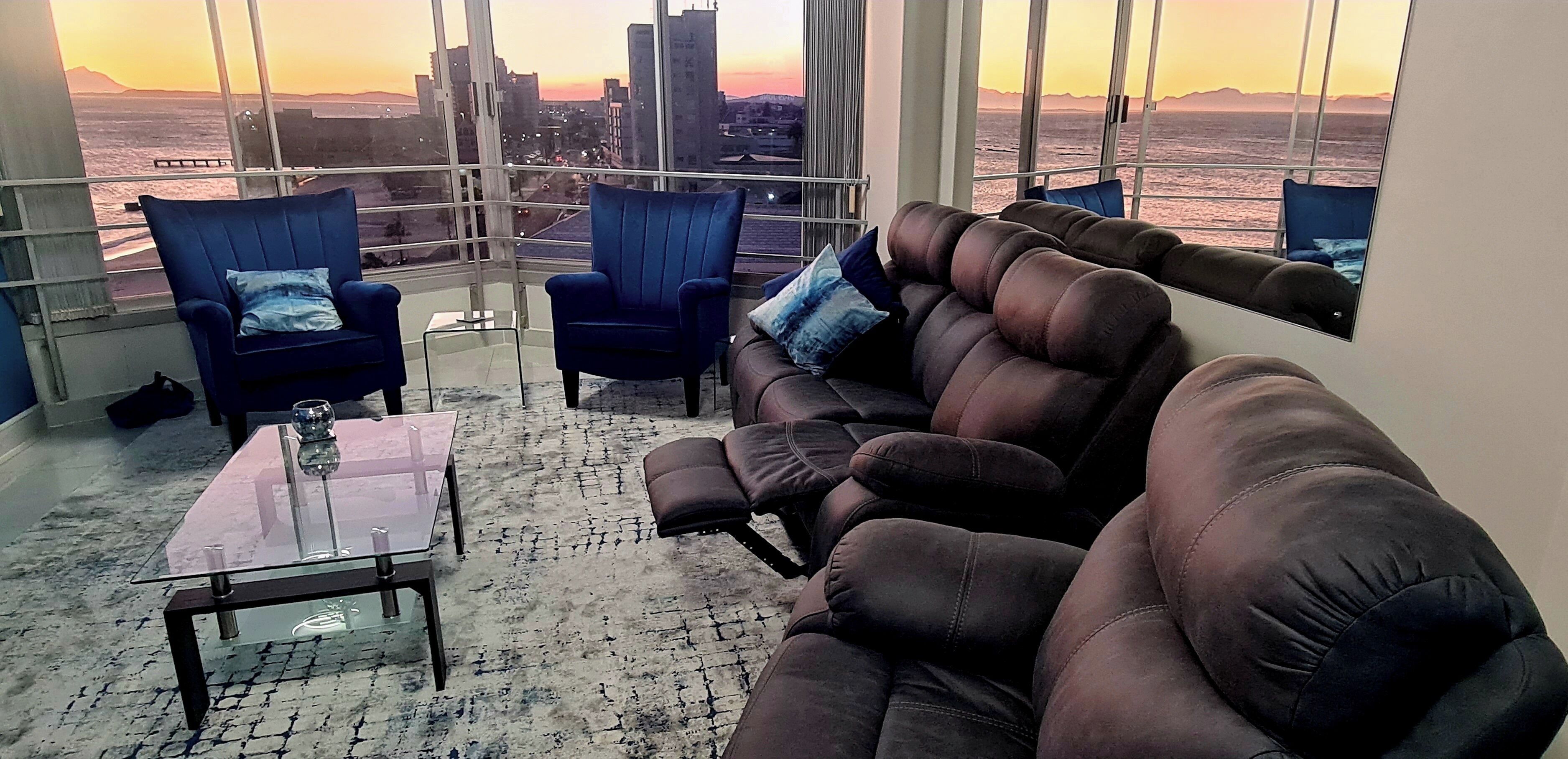 A modern living room with a leather sofa, two blue armchairs, and a glass coffee table. Large windows offer a view of the city skyline and a body of water during sunset.