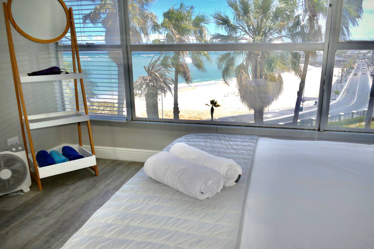 A bedroom with a large window, a bed, a wooden shelf, and a fan.