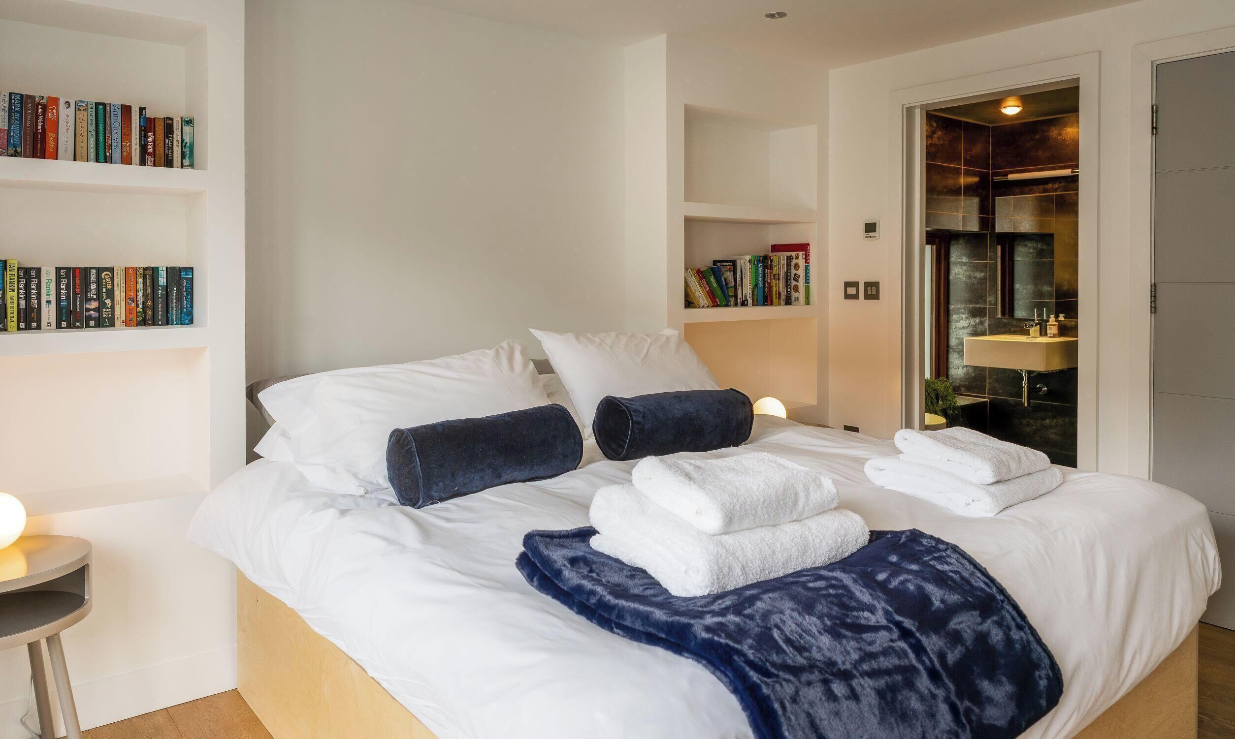 A neatly made bed with white linens, a dark blue throw blanket, and several folded white towels. There are bookshelves with books on the left side of the room.