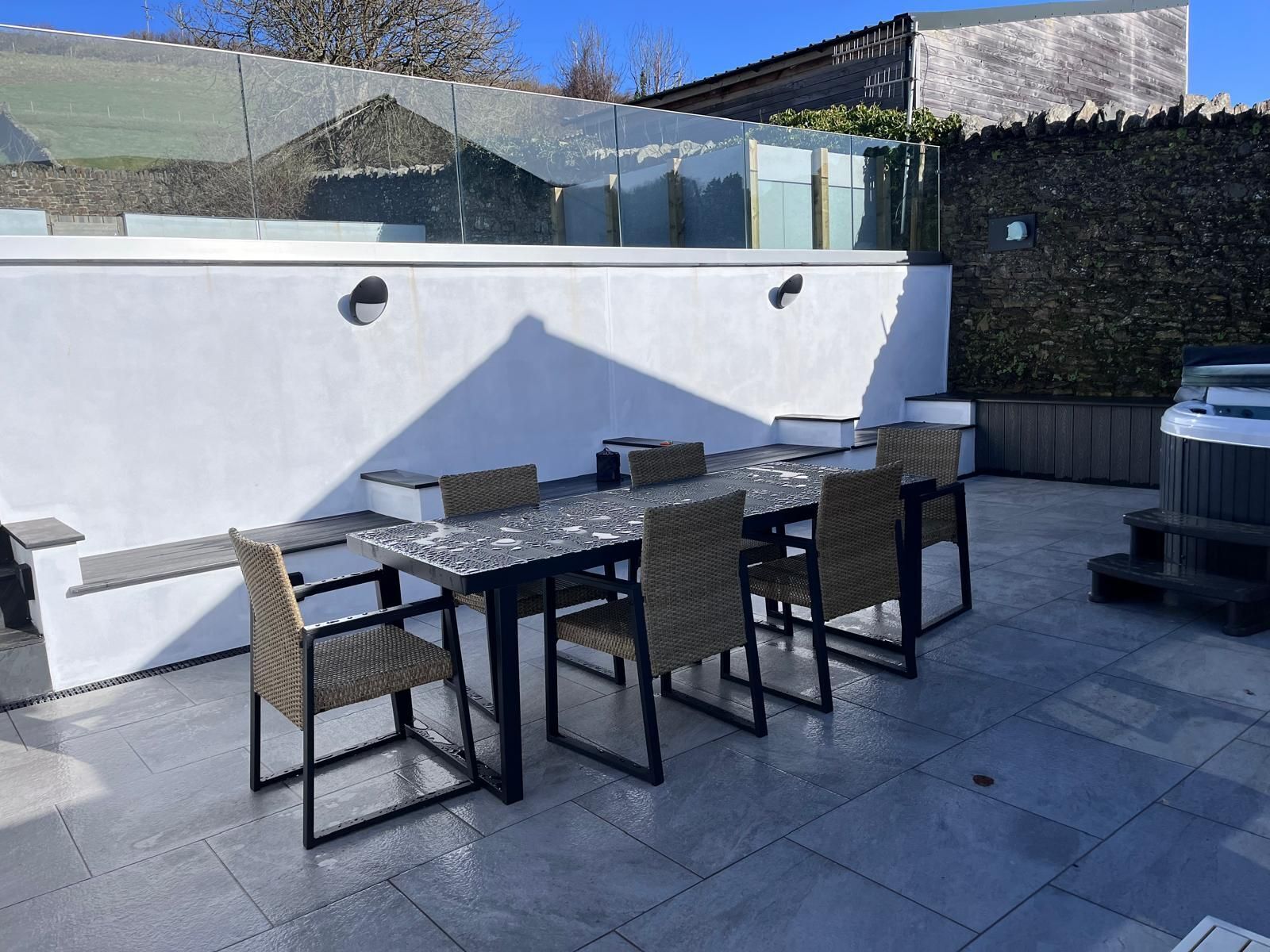 An outdoor patio with a rectangular table and four chairs, surrounded by stone walls and a glass enclosure reflecting the landscape.