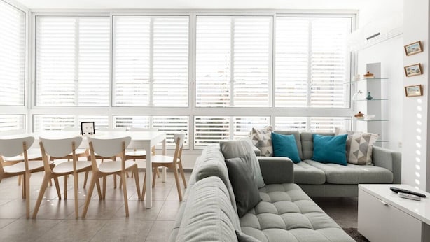 Ein modernes Wohnzimmer mit einem grauen Sofa, einem Esstisch mit Stühlen und einem großen Fenster mit Jalousien.