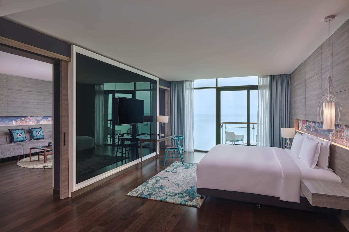 A modern hotel room with a large bed, a seating area with a sofa and chairs, a dining table, and a balcony with a view.