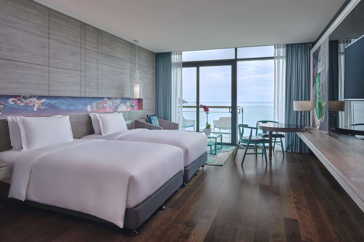 A modern hotel room with two beds, a balcony with a view of the sea, and a dining area with a table and chairs.