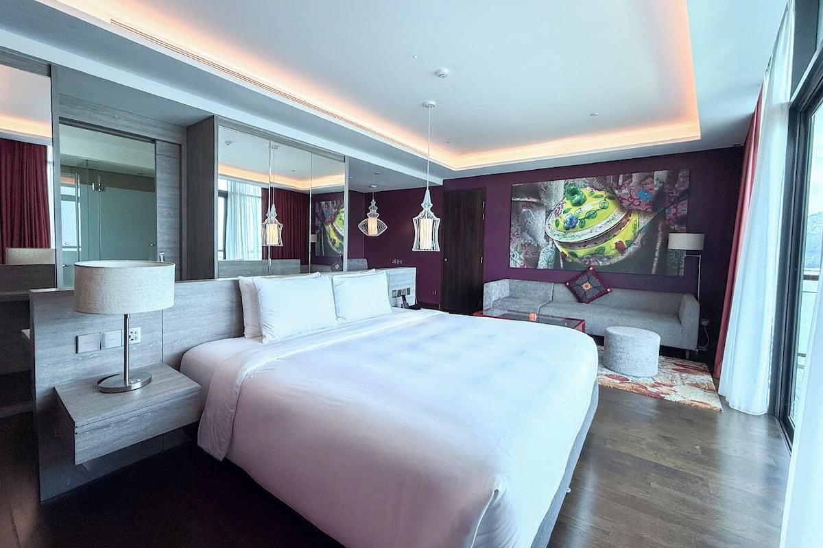 A modern hotel room with a large bed, a sofa, a desk with a lamp, and a large mirror.