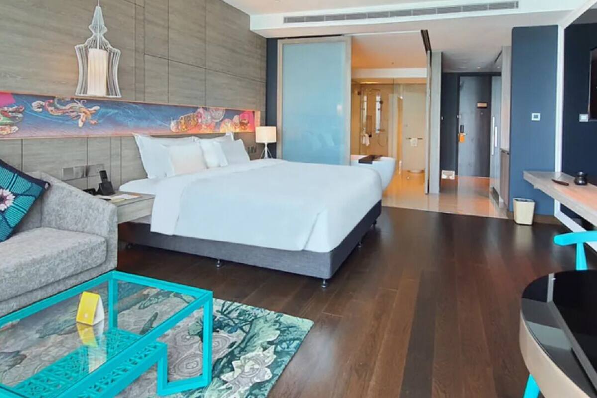 A modern hotel room with a large bed, a sofa, a glass coffee table, and a desk.