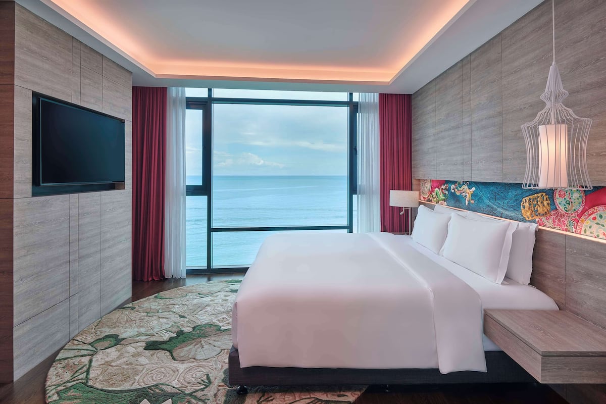 A hotel room with a large bed, a flat-screen TV mounted on the wall, a large window with a view of the sea, and a colorful artwork above the bed.
