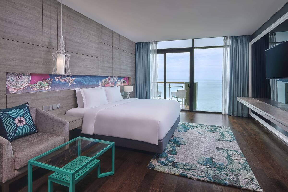 A hotel room with a bed, a sofa, a glass coffee table, a large window with a view of the sea, and a flat-screen TV.