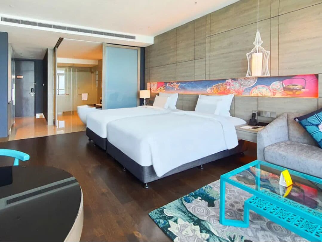 A modern hotel room with a large bed, a sofa, a glass coffee table, and a colorful abstract painting on the wall.