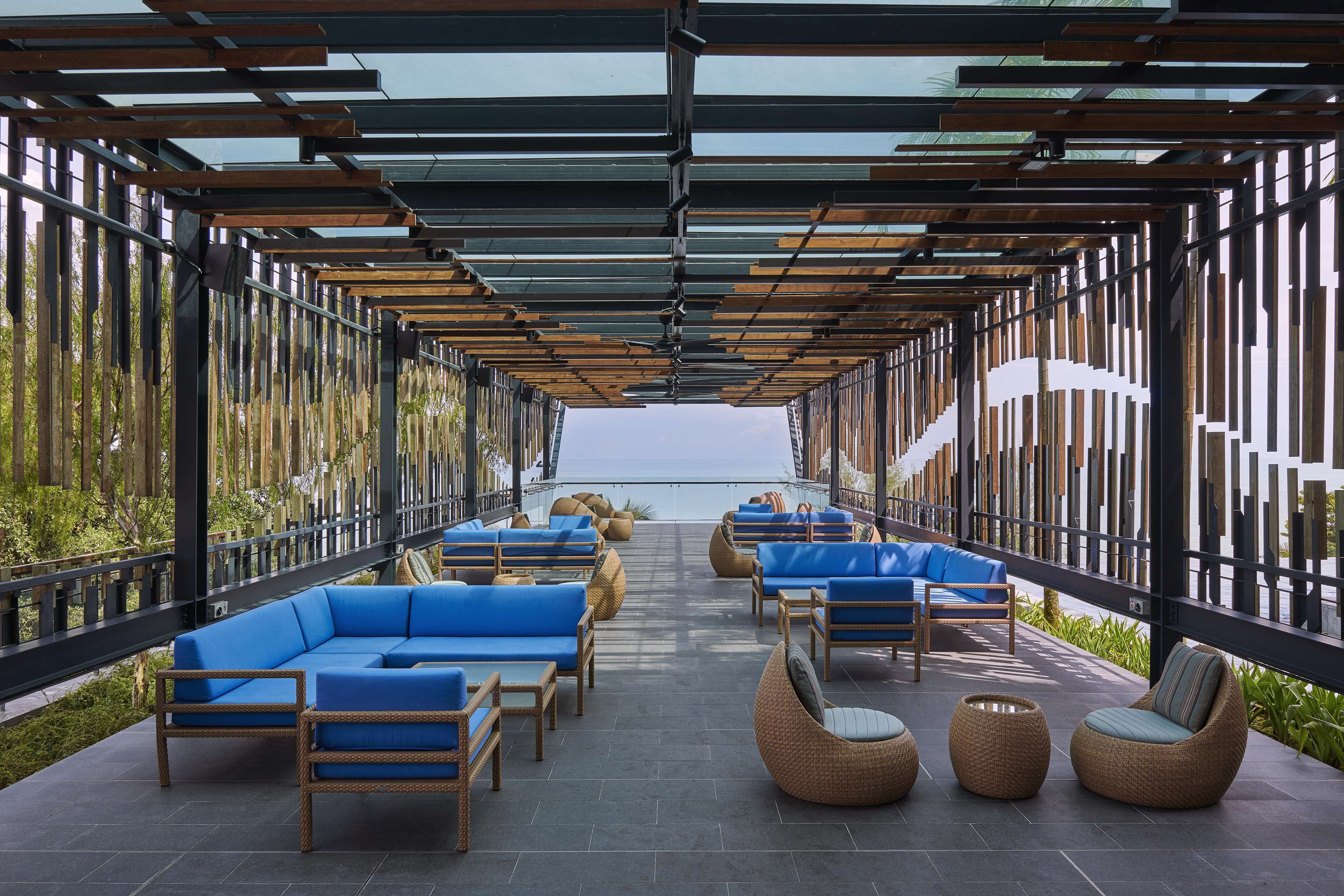 Een overdekt terras met blauwe, beklede banken en rieten stoelen, met uitzicht op een wateroppervlak.