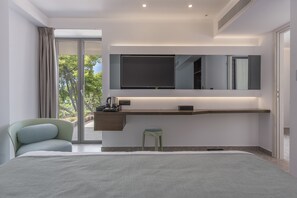 Quarto de hotel moderno com uma cama grande, uma escrivaninha com televisão de tela plana, um banquinho pequeno e uma janela ampla com vista para árvores.