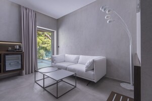 A modern living room with a white sofa, a glass-top coffee table, a floor lamp, and a view of greenery outside.