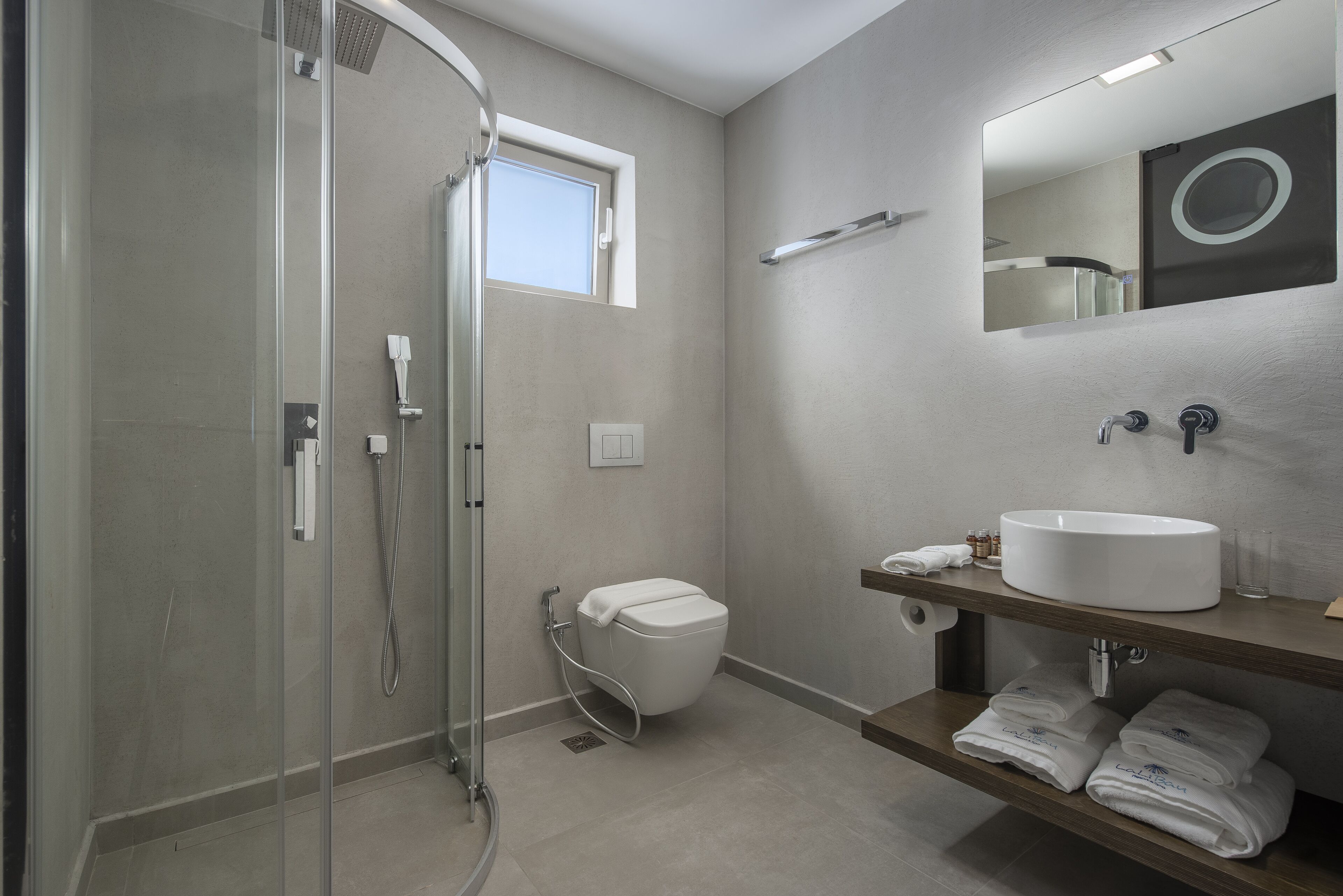 Un bagno moderno con cabina doccia in vetro, un water bianco, un lavabo con specchio e asciugamani piegati ordinatamente su uno scaffale.