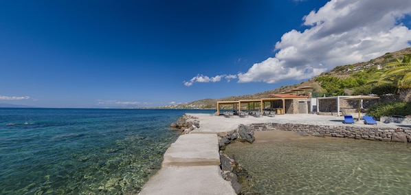 A coastal hotel with a clear view of the sea, a stone pathway leading to the beach, and a shaded outdoor seating area.