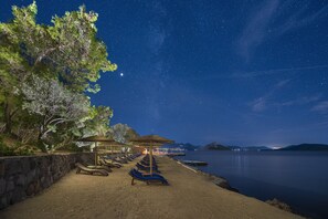 A beach with lounge chairs and umbrellas, a stone wall, and a calm lake under a starry night sky.