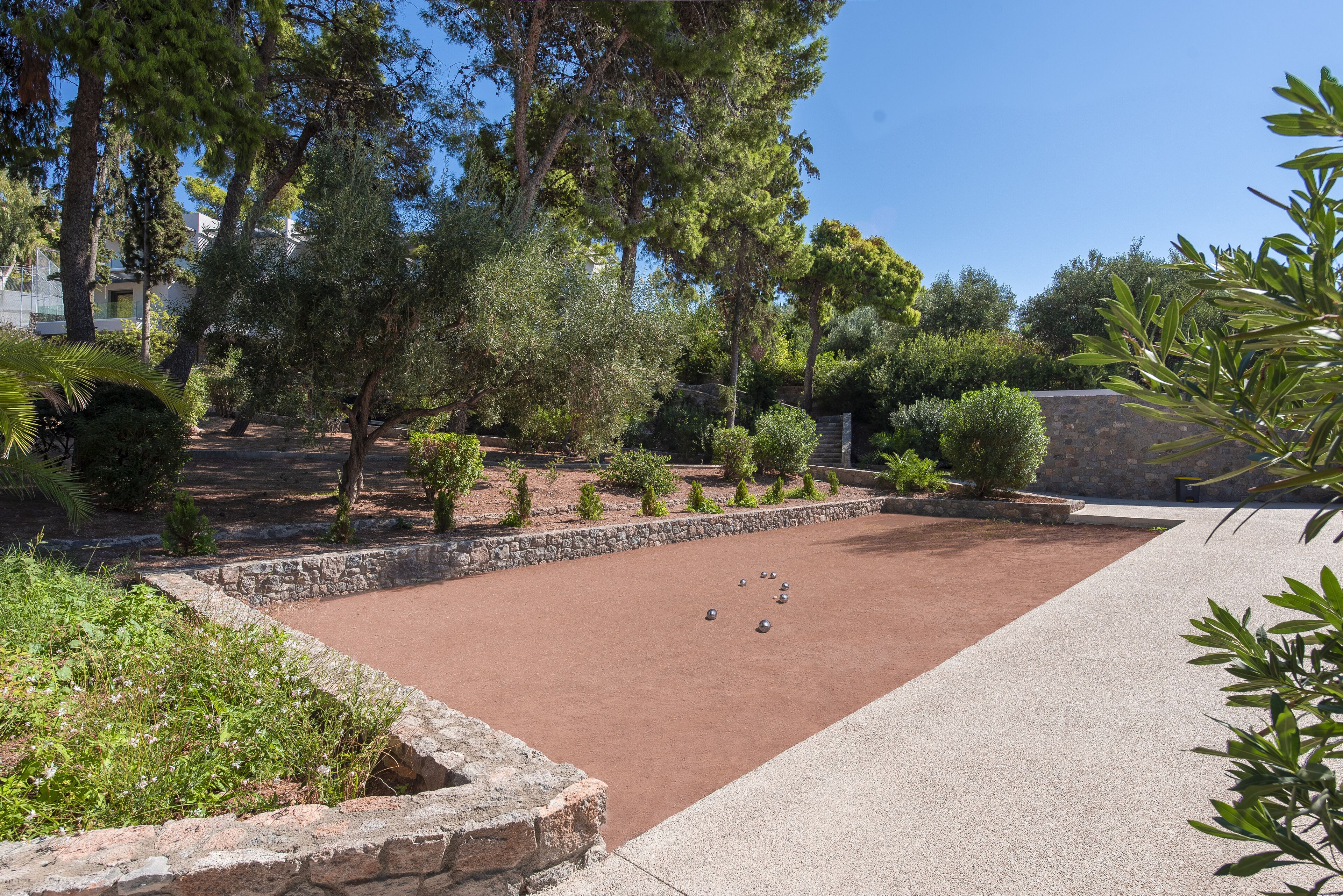 Un patio con superficie rojizo-marrón, rodeado de muros de piedra y varios árboles.